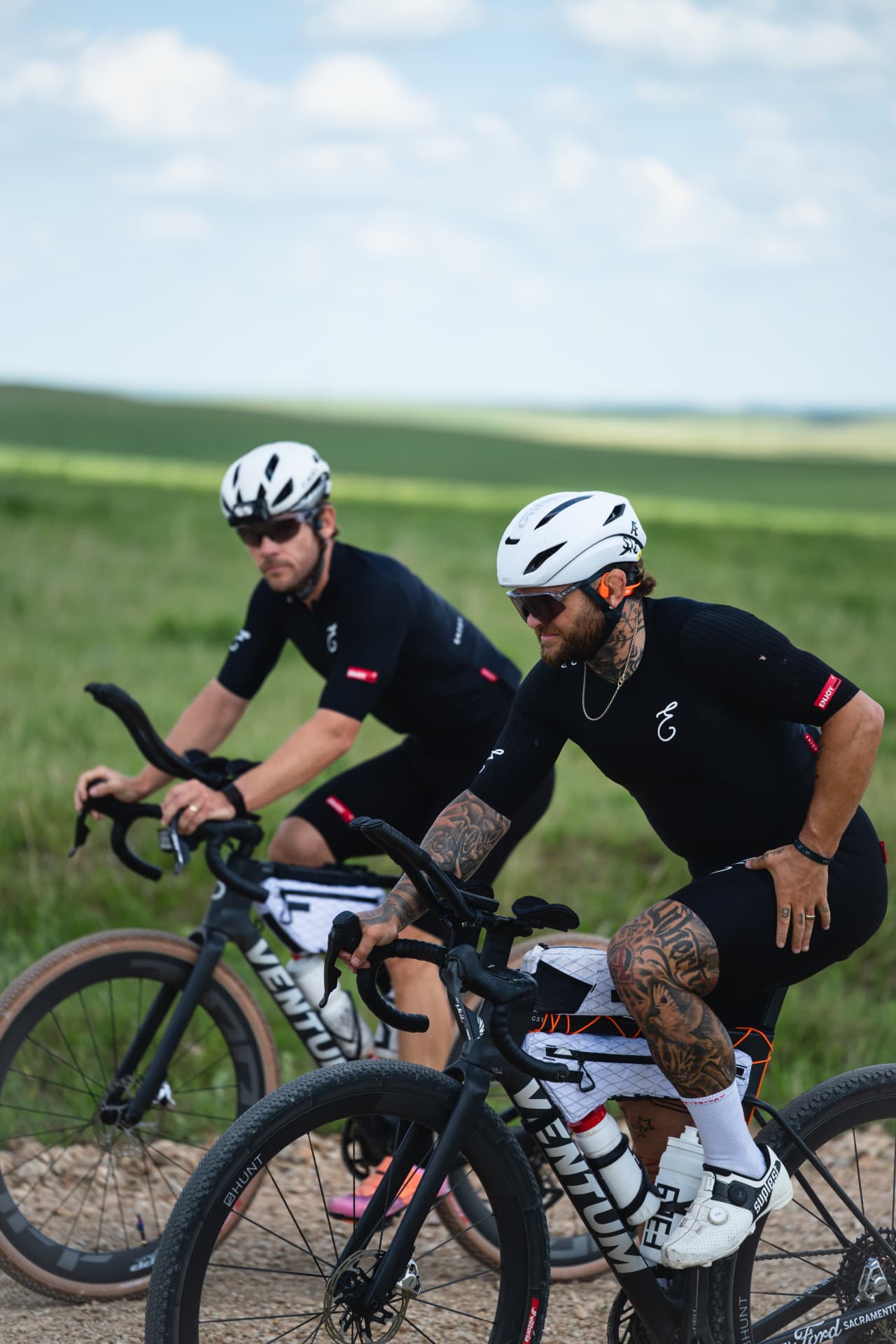Two riders in black Enjoy kit on a gravel road.
